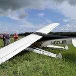 Small white light aircraft down in a grassy field with several men examining the scene; a red tow truck is in the background.