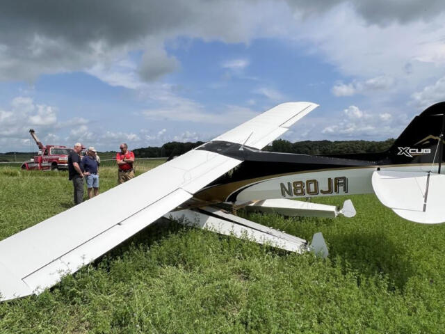 Small white light aircraft down in a grassy field with several men examining the scene; a red tow truck is in the background.