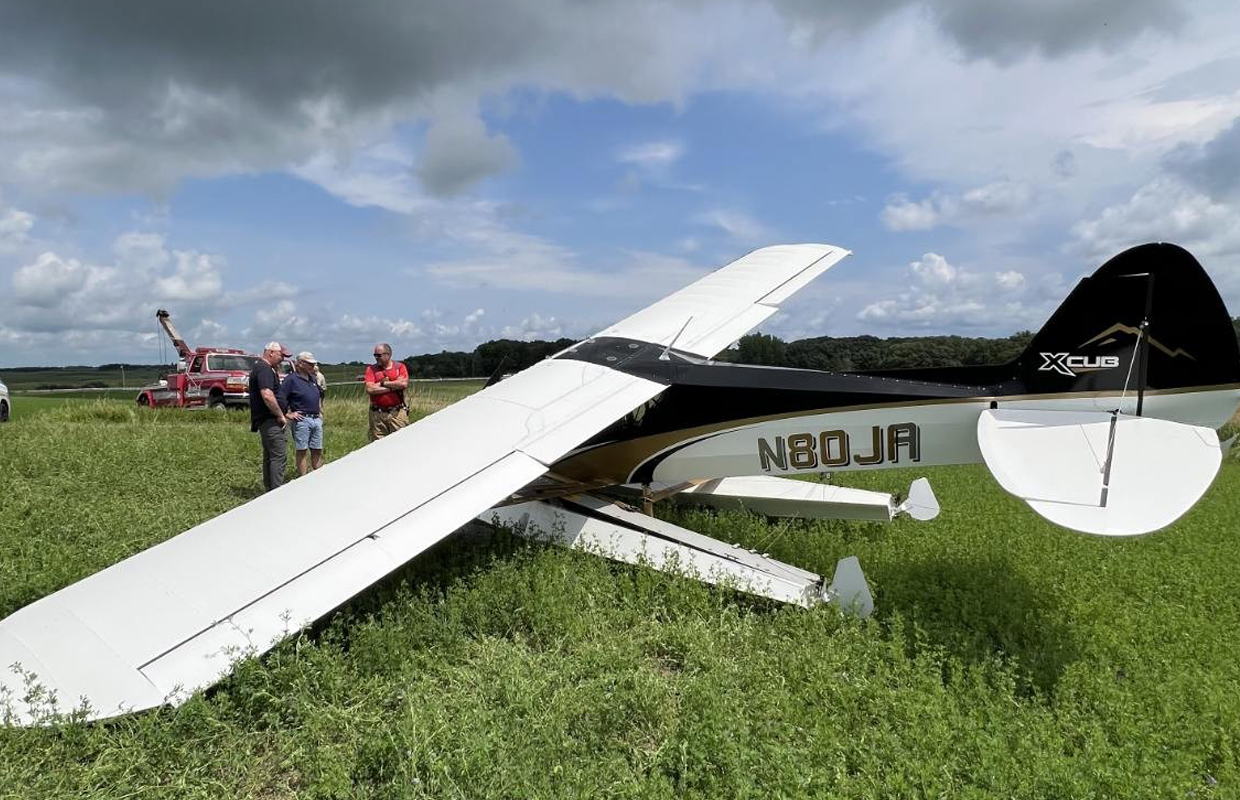 Small white light aircraft down in a grassy field with several men examining the scene; a red tow truck is in the background.