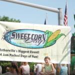Group of people at a festival holding a banner that reads 'Sweet Corn Day' with corn graphics, promoting Estherville, IA and '4 Jam Packed Days of Fun!'