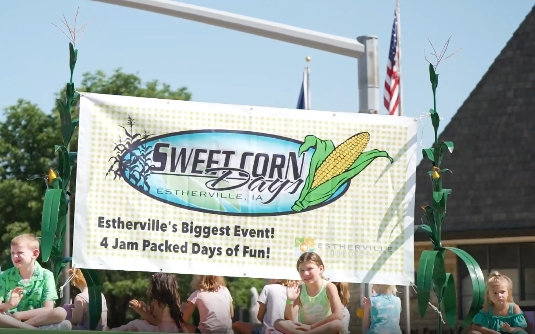 Group of people at a festival holding a banner that reads 'Sweet Corn Day' with corn graphics, promoting Estherville, IA and '4 Jam Packed Days of Fun!'