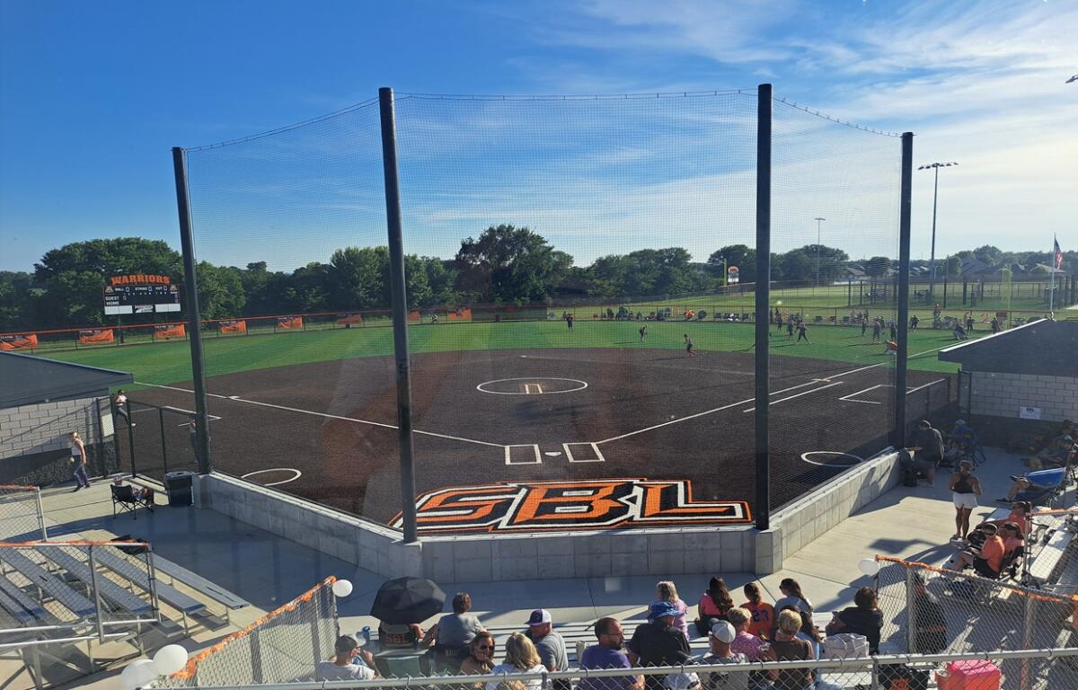 Baseball field with protective netting around home plate and bleachers full of spectators under a clear blue sky.