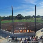 Baseball field with protective netting around home plate and bleachers full of spectators under a clear blue sky.