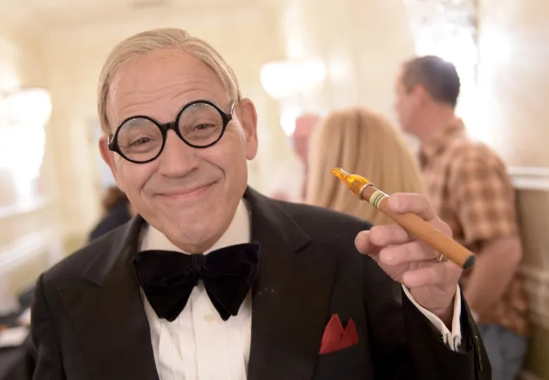 Smiling older man in a tuxedo with bow tie and red pocket square, holding a party horn toward the camera.